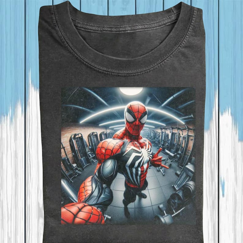 May include: Dark gray t-shirt featuring a graphic of Spider-Man in a gym setting. The superhero is in a red and black suit, striking a pose. The background shows gym equipment and bright lights.