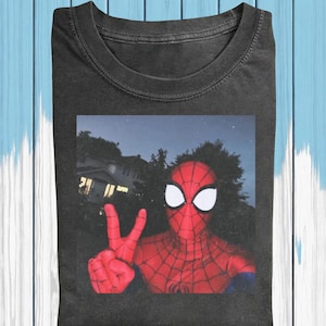 May include: Dark gray t-shirt featuring a selfie-style image of Spider-Man making a peace sign. The superhero is in full costume, with a red and black webbed suit and white eye lenses. The background shows a house and trees under a night sky.