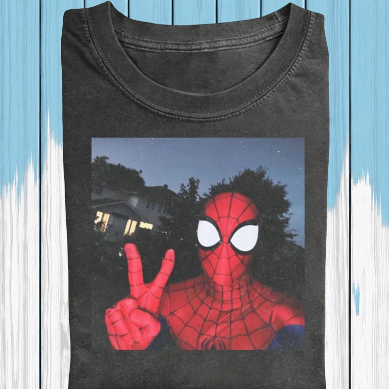 May include: Dark gray t-shirt featuring a selfie-style image of Spider-Man making a peace sign. The superhero is in full costume, with a red and black webbed suit and white eye lenses. The background shows a house and trees under a night sky.