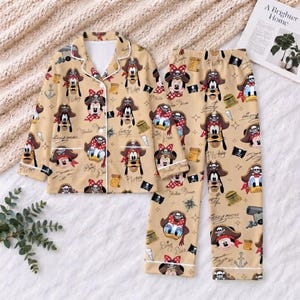 May include: A beige pyjama set featuring a long-sleeved button-down shirt and matching trousers. The fabric is covered in a repeating pattern of cartoon characters wearing pirate hats, along with small illustrations of treasure chests and anchors.