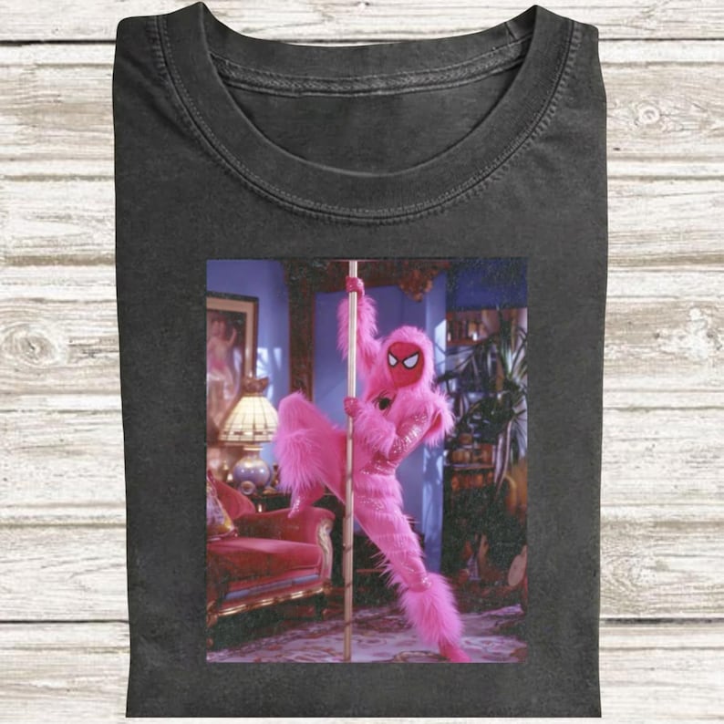 May include: A charcoal gray crew neck t-shirt featuring a graphic print of a fuzzy pink character wearing a red mask with white eye lenses, posed on a vertical pole. The character has long, shaggy pink fur and is positioned in a room setting.
