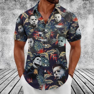 Puede incluir: Una camisa de manga corta con botones sobre un fondo azul marino oscuro, que presenta un patrón repetitivo de máscaras de Michael Myers intercaladas con hojas tropicales coloridas en tonos naranja, amarillo y verde por toda la prenda.