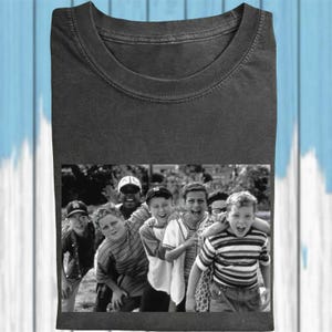 May include: Dark gray t-shirt featuring a black and white image of a group of boys. The image shows the boys smiling and laughing. The t-shirt has a crew neck and short sleeves.