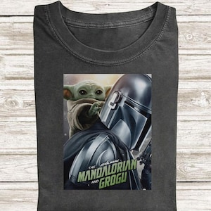 May include: Dark gray t-shirt featuring a graphic of The Mandalorian and Grogu from Star Wars. The image shows the Mandalorian's helmet and Grogu, with the text "The Mandalorian and Grogu" in green and white.