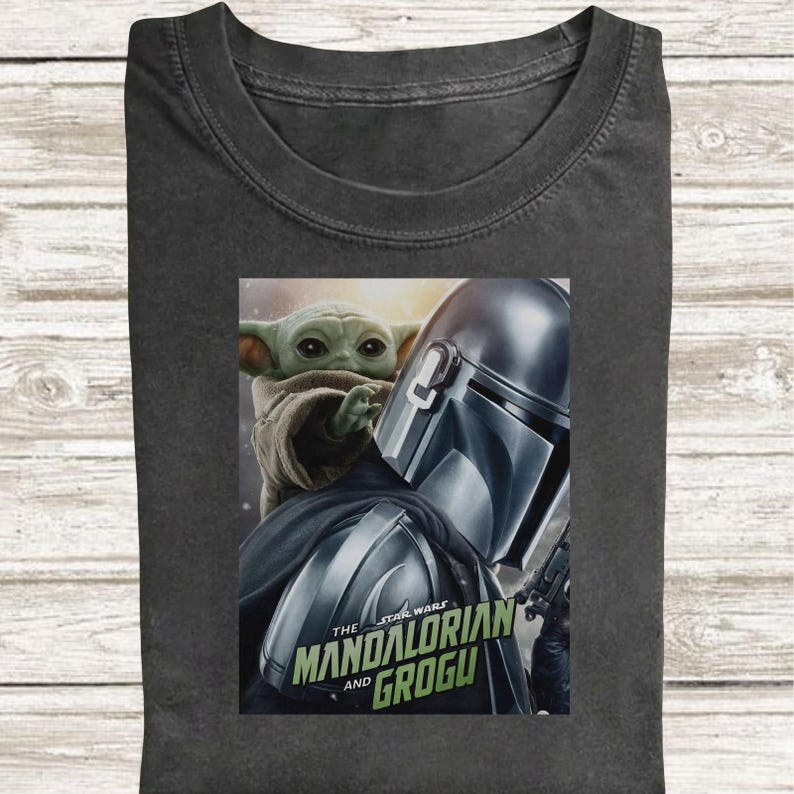 May include: Dark gray t-shirt featuring a graphic of The Mandalorian and Grogu from Star Wars. The image shows the Mandalorian's helmet and Grogu, with the text "The Mandalorian and Grogu" in green and white.