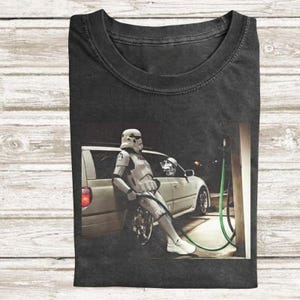 May include: Dark gray t-shirt featuring a graphic of a Stormtrooper leaning against a car at a gas pump. The Stormtrooper is wearing a white helmet and armor. The car is light-colored, and the gas pump hose is green.