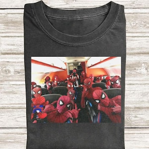 May include: A charcoal gray t-shirt featuring a graphic print of an airplane cabin filled with numerous individuals wearing red and blue Spider-Man costumes. The scene depicts a crowded interior with multiple figures in masks sitting in rows.