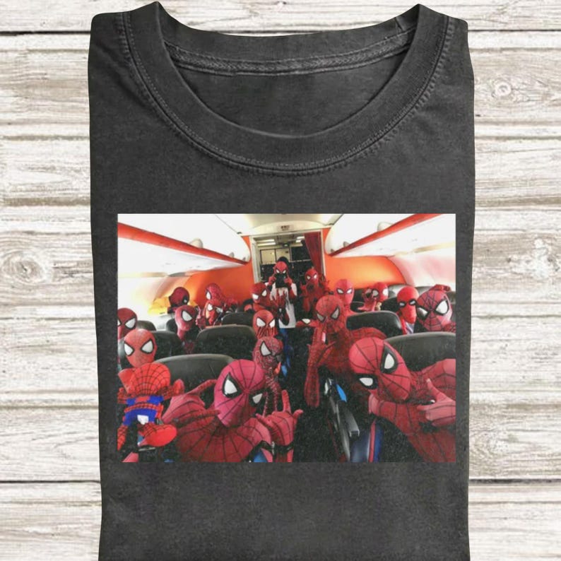 May include: A charcoal gray t-shirt featuring a graphic print of an airplane cabin filled with numerous individuals wearing red and blue Spider-Man costumes. The scene depicts a crowded interior with multiple figures in masks sitting in rows.
