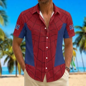 K&ouml;nnte beinhalten: Rotes und blaues kurz&auml;rmeliges Hemd mit Spinnennetz-Design. Das Hemd hat blaue Eins&auml;tze an den Seiten und die Buchstaben "IN" und "AL" auf den &Auml;rmeln. Das Hemd wird an einem Strand getragen.