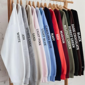 May include: A rack displays a variety of sweatshirts in various colors, including white, ash, sports grey, sand, light pink, light blue, Carolina blue, navy, red, maroon, military green, forest green, dark chocolate, and black. Each sweatshirt is labeled with its color.