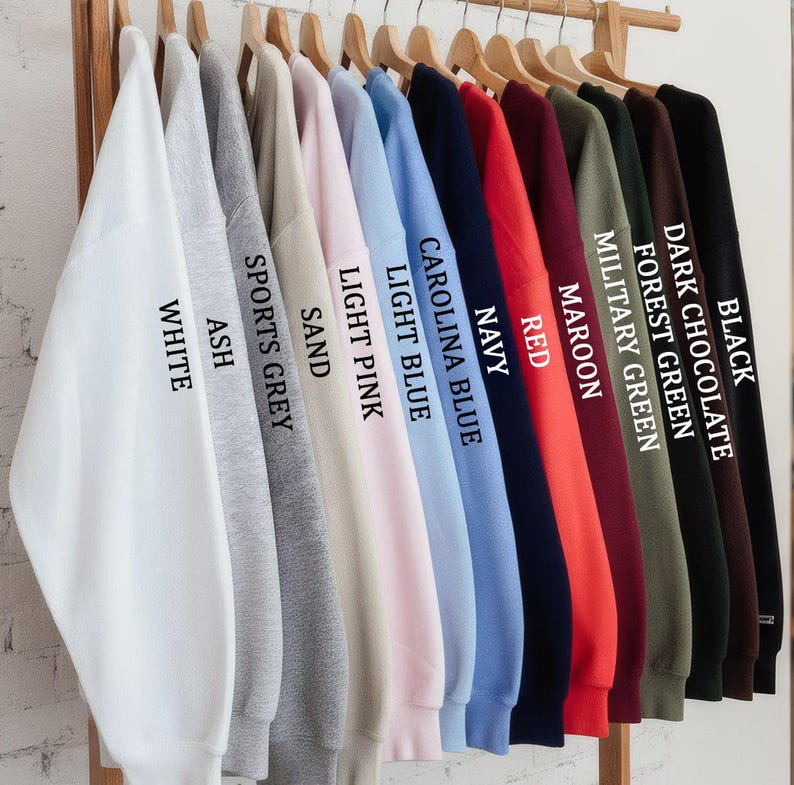 May include: A rack displays a variety of sweatshirts in various colors, including white, ash, sports grey, sand, light pink, light blue, Carolina blue, navy, red, maroon, military green, forest green, dark chocolate, and black. Each sweatshirt is labeled with its color.