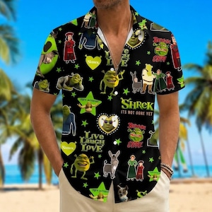 Könnte beinhalten: Ein schwarzes kurzärmeliges Hemd mit Knopfleiste und einem wiederkehrenden Muster aus Shrek-Figuren, grünen Sternen und Herzen. Der Text umfasst "Shrek", "It's Not Ogre Yet", "Live Laugh Love" und "Beware Ogre". Das Design zeigt Shrek in verschiedenen Posen.