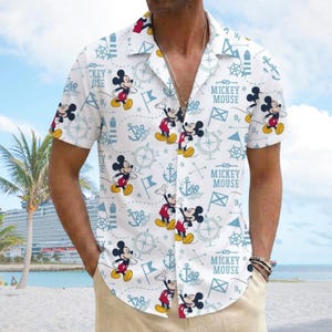 May include: White button-down shirt with a repeating pattern of Mickey Mouse, anchors, compasses, and flags. The shirt features short sleeves and the text "MICKEY MOUSE" and "CKEY USE". The shirt is worn on a beach.