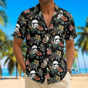 Puede incluir: Una camisa de manga corta con botones sobre un fondo negro, con un patrón repetitivo de cascos de Stormtrooper blancos, cazas estelares oscuros, flores de hibisco tropicales de colores y hojas de palmera verdes por toda la tela.