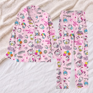 Può includere: Un set di pigiama rosa chiaro composto da una camicia a maniche lunghe con bottoni e pantaloni abbinati. Il tessuto presenta un motivo ripetuto di personaggi Hello Kitty, palloncini colorati, palle da discoteca e il testo Be the best version of yourself.