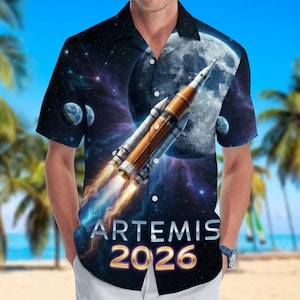 May include: Short-sleeved button-down shirt with a space-themed design. The shirt features a rocket blasting off towards the moon, with the text "ARTEMIS 2026" in silver and gold. The background includes planets and a starry sky.