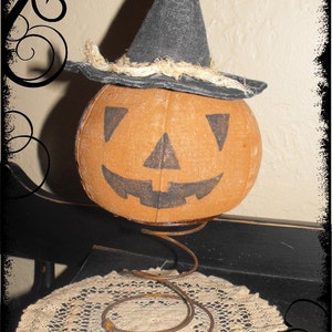 May include: A handmade fabric pumpkin with a black witch hat and a bell on top. The pumpkin is sitting on a metal spring and a white crocheted doily.