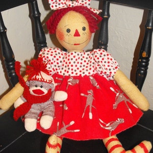 Peut inclure: Une poupée de chiffon vêtue d'une robe à pois rouges et blancs et de bas rayés rouges et blancs est assise sur une chaise noire. La poupée tient un singe en chaussette gris avec une écharpe rouge.