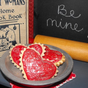 May include: A vintage cookbook and heart-shaped cookies on a metal plate. The cookies are red with silver glitter, and the plate sits on a red and white cloth. A rolling pin and a chalkboard with "Be mine!" are in the background.