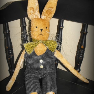 May include: A plush bunny rabbit wearing a black and white checkered vest and a yellow bow tie. The bunny is sitting on a black wooden chair.