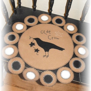Peut inclure: Un tapis en feutre brun avec un motif de corbeau noir et le texte "Olde Crow". Le tapis est entouré d'un cercle de plus petits cercles en feutre noir et blanc.
