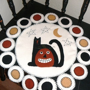 May include: A white felt Halloween doormat with a black cat sitting on a jack-o'-lantern. The doormat is surrounded by a circle of felt circles in black, brown, and tan.