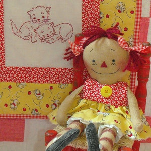 May include: A rag doll with red hair and a yellow dress sits in a red wooden chair. The doll is smiling and has a red and white polka dot dress. The chair has a woven straw seat.