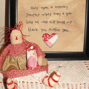 May include: A framed piece of art with a handwritten message that reads "Once upon a memory Someone wiped away a tear held me close and loved me thank you. Mother Dear."  A red and white patchwork heart is in the lower centre of the frame. A red and white rag doll with a red and white dress and red and white striped legs is sitting in front of the frame.