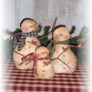 May include: Three handcrafted snowman figures with twig arms and carrot noses. Each snowman wears a plaid scarf and has a heart-shaped patch. The snowmen are arranged on a red and cream checkered surface, with evergreen branches in the background.