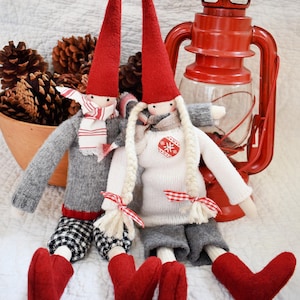 Op de afbeelding: Twee handgemaakte kerstkabouters met rode puntmutsen, grijze truien en rode hartvormige laarzen. Eén kabouter draagt een sjaal, de ander heeft vlechten. Een rode lantaarn en dennenappels maken de scène af.