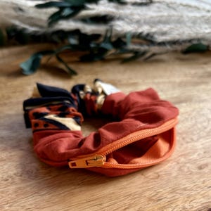 May include: A rust-colored fabric scrunchie featuring a hidden zipper compartment. One side of the scrunchie displays a patterned section with black, cream, and rust-colored geometric shapes, while the rest is solid rust fabric with an orange zipper.