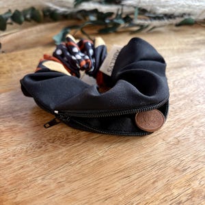 May include: A black fabric scrunchie featuring a hidden zipper compartment along the outer edge. The scrunchie has a small white tag with the text ABELYS and contains a copper-colored coin inside the zippered pocket. The fabric has a pattern.