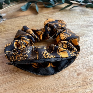 Peut inclure: Un chouchou en tissu arborant un motif floral noir et jaune moutarde avec une poche zippée noire intégrée. Cet accessoire de coiffure circulaire présente une texture froncée et une fermeture éclair fonctionnelle sur le bord extérieur.