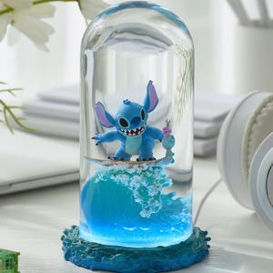 May include: A blue Stitch figurine surfing a wave inside a glass dome. The base of the dome is a textured blue, and the wave is illuminated with blue light. A smaller figurine is next to Stitch.