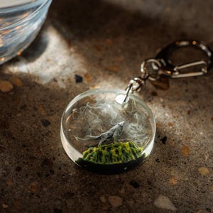 May include: A round keychain with a clear resin pendant. Inside the pendant is a gray wolf figurine on a green and black base, with white wisps. The keychain has a silver-colored metal clasp.