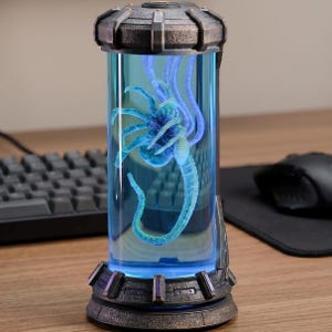 May include: A futuristic display piece, cylindrical in shape, showcasing a blue alien creature within a transparent tube. The creature has a detailed, skeletal form with multiple limbs and a long tail. The tube is framed by a dark, metallic base and top, with blue internal lighting.
