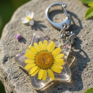 Può includere: Un portachiavi a forma di stella con un fiore giallo racchiuso in resina trasparente. Il portachiavi ha un moschettone argentato e un piccolo ciondolo a forma di ape. Glitter rosa accentuano la resina, e i petali gialli e il centro marrone del fiore sono visibili.