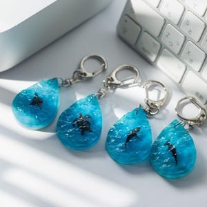 May include: Four teardrop-shaped keychains with a blue ocean design and a sea turtle or shark embedded within. Each keychain has a silver-colored clasp. The keychains are displayed near a white keyboard.
