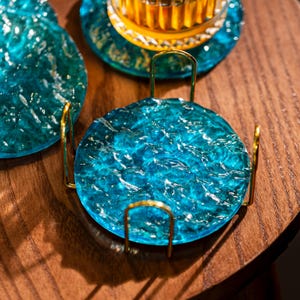 May include: A set of three decorative coasters. The coasters are a vibrant turquoise blue with a textured, water-like appearance. Each coaster sits in a gold-colored metal holder. The coasters are on a dark wood surface.