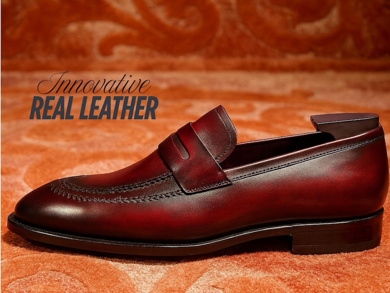 May include: A burgundy leather loafer with a classic design. The shoe features a stitched vamp, a strap across the instep, and a dark sole. The text "Innovative REAL LEATHER" is displayed in the image.