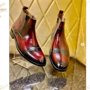 May include: A pair of leather Chelsea boots with a gradient finish, transitioning from burgundy to brown. The boots have a side zipper and are displayed on a clear stand, showcasing the craftsmanship and design.