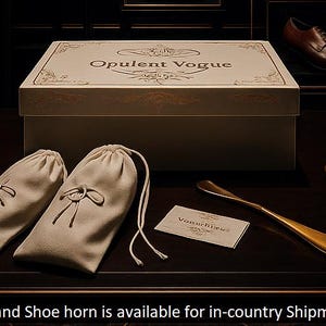 May include: A beige box with the text "Opulent Vogue" sits on a dark surface, accompanied by two beige pouches, a business card, and two gold-colored shoe horns. A pair of brown shoes is visible in the background.