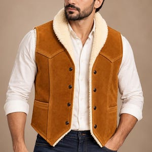 May include: A tan suede vest featuring a cream-colored faux shearling lining, a wide shawl collar, and a front button closure with four dark metal buttons. The vest includes two front pockets and western-style yoke detailing on the chest.
