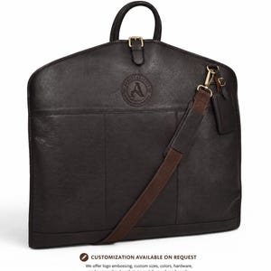 May include: A dark brown leather garment bag featuring a top handle, a brass buckle closure, and an adjustable shoulder strap. The front displays a circular embossed logo with the letter A and the text The Abbott Leather Co. Handcrafted in USA.