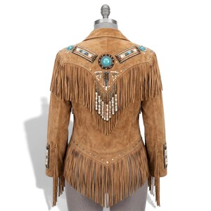 May include: A tan suede jacket featuring long fringe detailing on the back and sleeves. The back is decorated with a central turquoise stone, intricate beadwork, and metallic studs arranged in a V-shaped pattern across the shoulders.