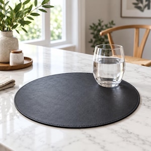 May include: A circular black placemat with a smooth, leather-like texture and stitched edges. The placemat is positioned on a white marble surface, with a clear glass of water resting on its right side, showcasing a clean and functional design.