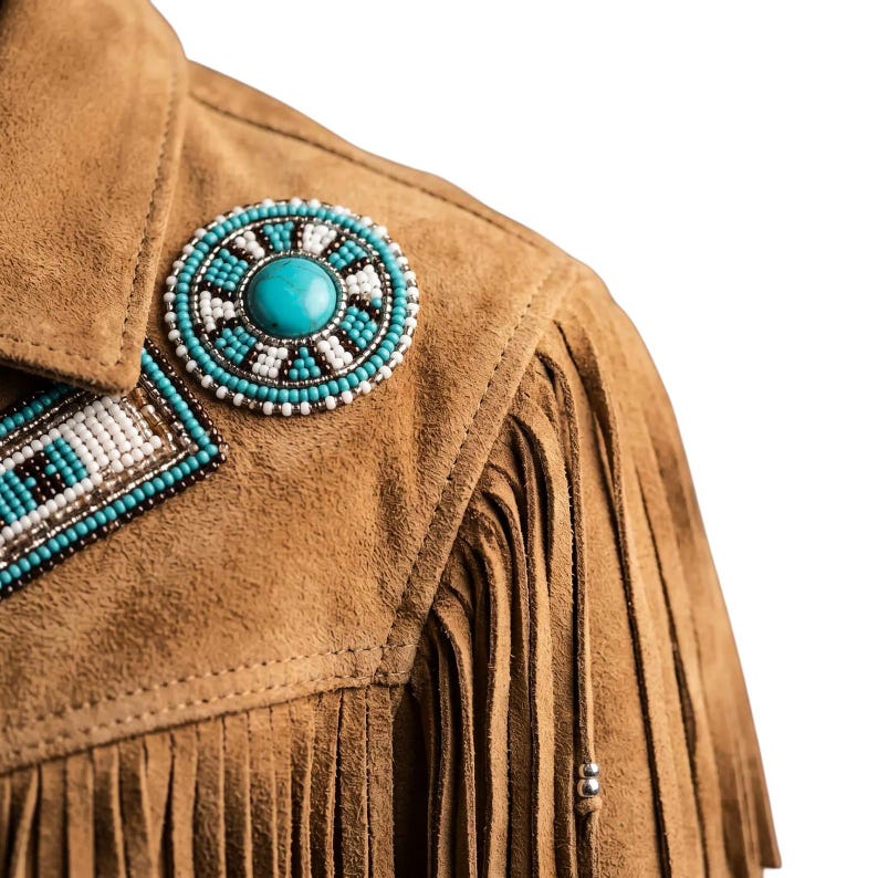 May include: A tan suede jacket featuring long fringe detailing along the shoulder and chest. The garment is embellished with a circular beaded patch containing a central turquoise stone, surrounded by white, blue, and black seed beads.
