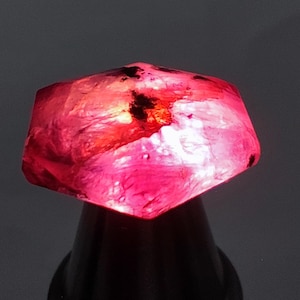 May include: A raw, faceted gemstone in shades of pink and red. The gem is lit, revealing its internal structure and colour variations. The stone is mounted on a dark base, set against a neutral background.