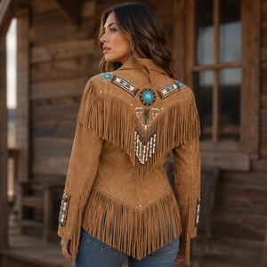 May include: A tan suede jacket featuring long fringe detailing along the back yoke and hem. The back is embellished with a central turquoise stone, intricate beadwork in shades of blue, white, and brown, and dangling beaded fringe accents.