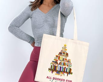 May include: Cream-colored tote bag featuring a Christmas tree design made of stacked books. The design includes a gold star on top and the text "ALL BOOKED FOR Christmas". The bag has long handles.